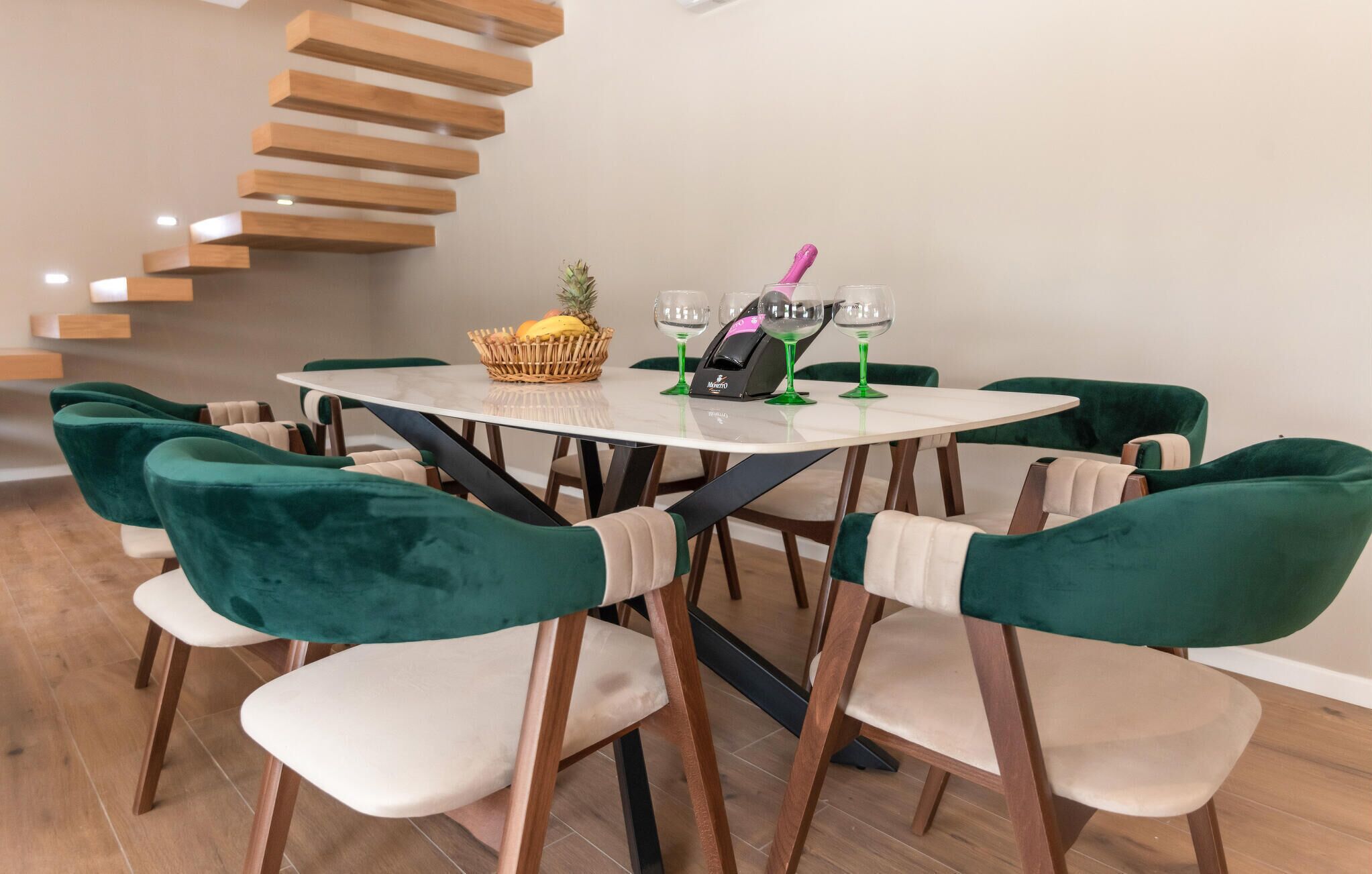 A dining area with a round table, green upholstered chairs, a wooden staircase, and a fruit basket.