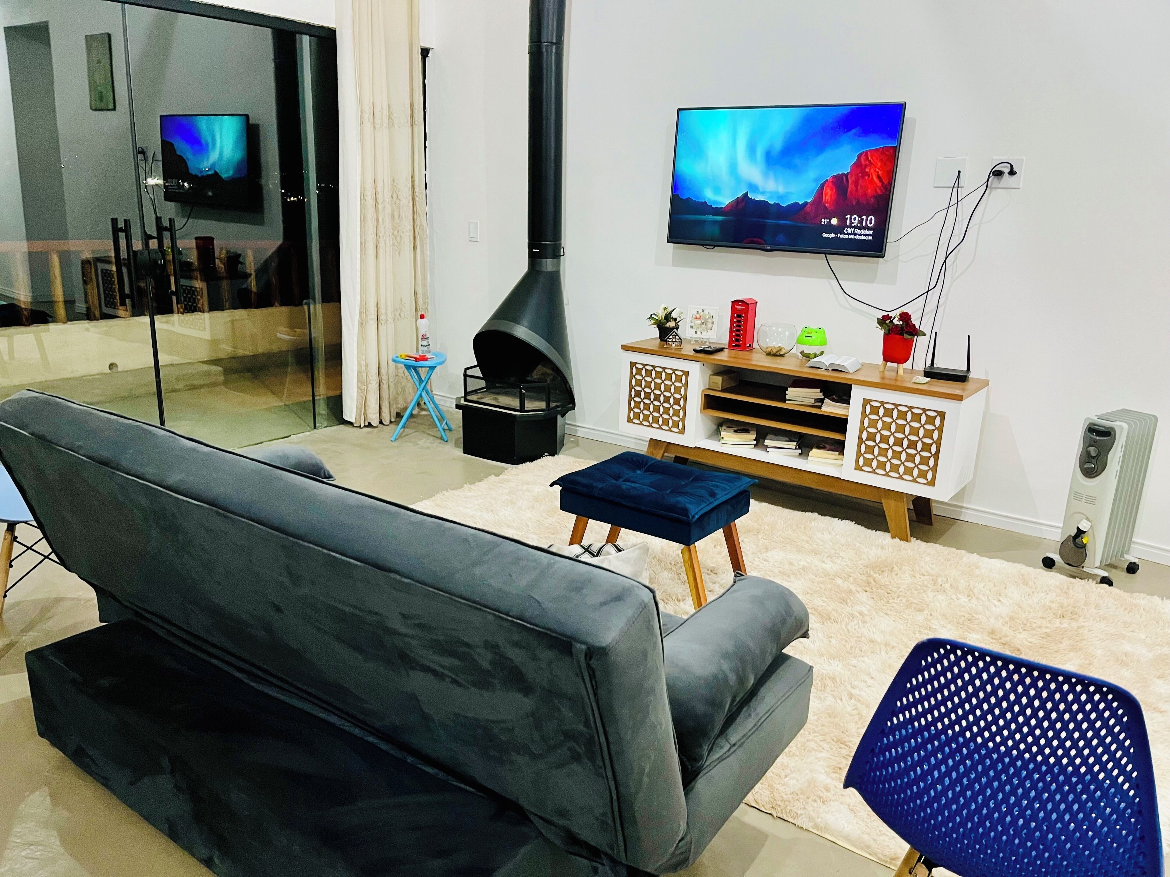 A modern living room with a grey sofa, a blue ottoman, a TV on the wall, a black fireplace, and a white fluffy rug.