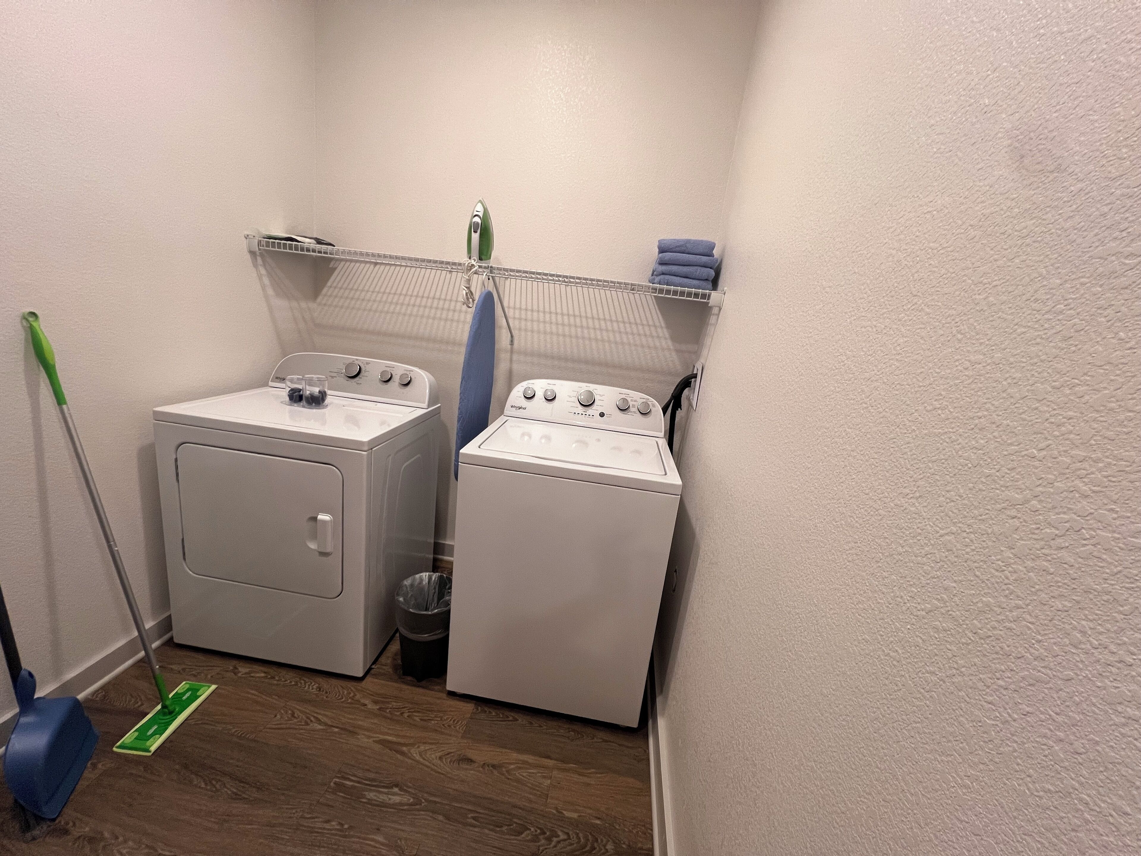 A laundry area with two washing machines, a drying rack, a mop, and a trash bin.