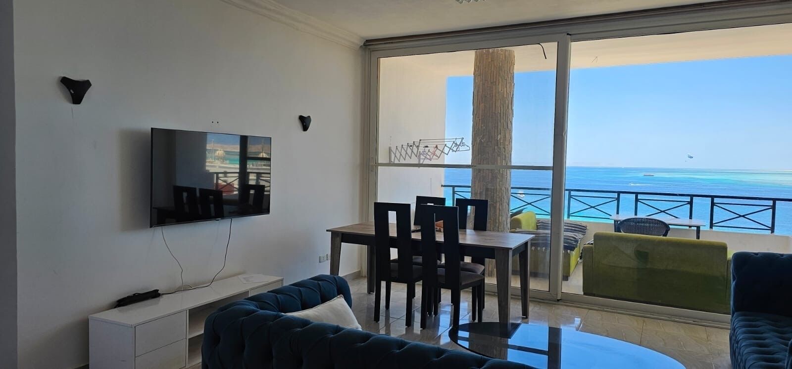 A balcony with a view of the sea, a dining area with chairs and a table, a sofa, and a TV mounted on the wall.