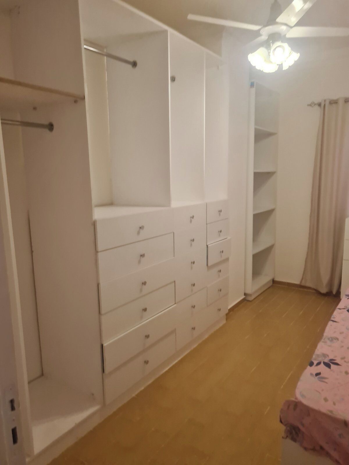 Ein Schlafzimmer mit Bett, Kleiderschrank mit Schubladen, Deckenventilator und einem Fenster mit Vorhängen.