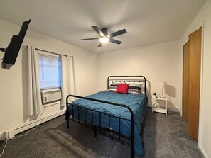 Une chambre à coucher comprenant un lit, un ventilateur de plafond, une table de chevet, une lampe et une fenêtre avec des rideaux.