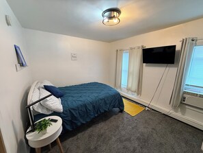 Un dormitorio con una cama, un televisor montado en la pared, una ventana con cortinas y una mesita con una planta.