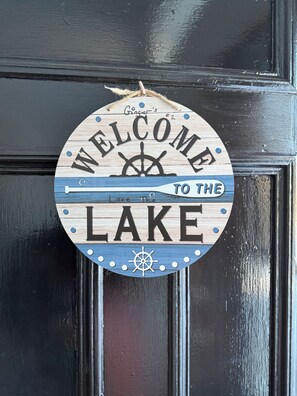 A circular wooden sign with a nautical theme, featuring the words "WELCOME TO THE LAKE" and a ship's wheel.