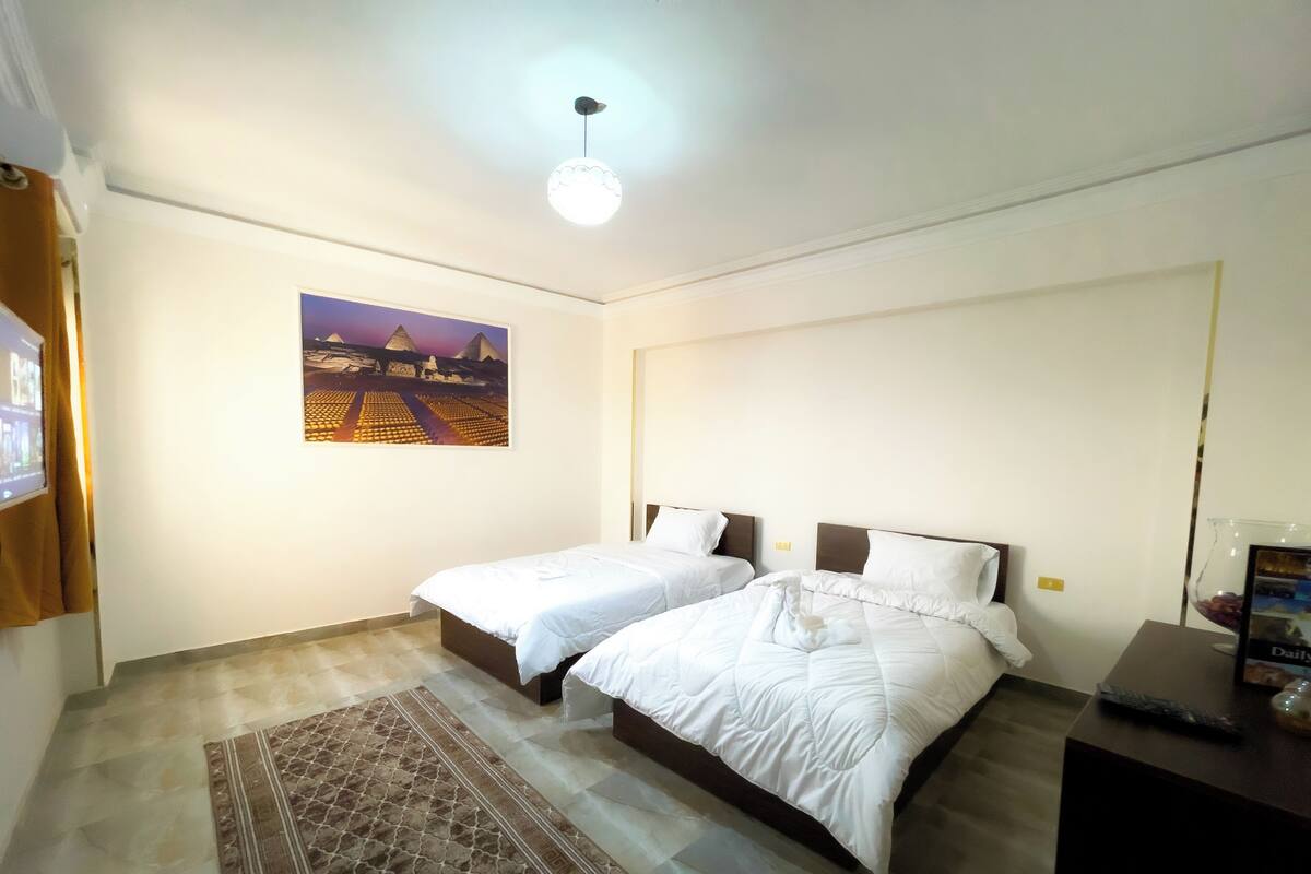 A hotel room with two beds, a rug, a painting of pyramids, and a desk with a keyboard.