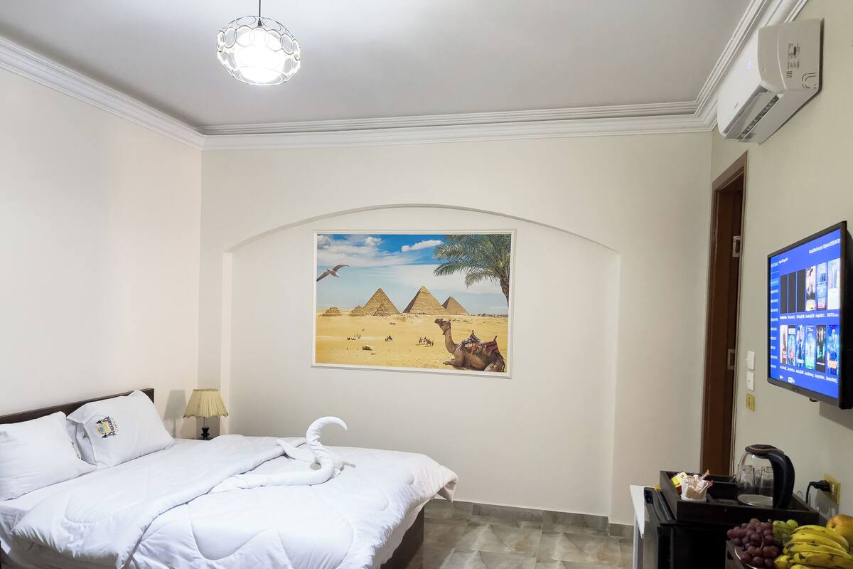 A hotel room with a bed, a wall-mounted TV, a painting of pyramids, and a fruit basket.