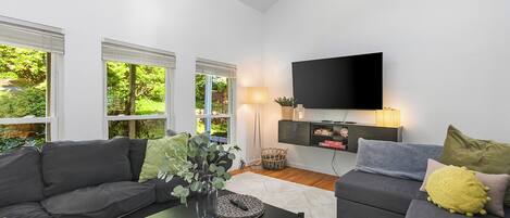 Ein modernes Wohnzimmer mit großem Flachbildfernseher, zwei Sofas, einem Couchtisch und einem großen Fenster mit Jalousien.