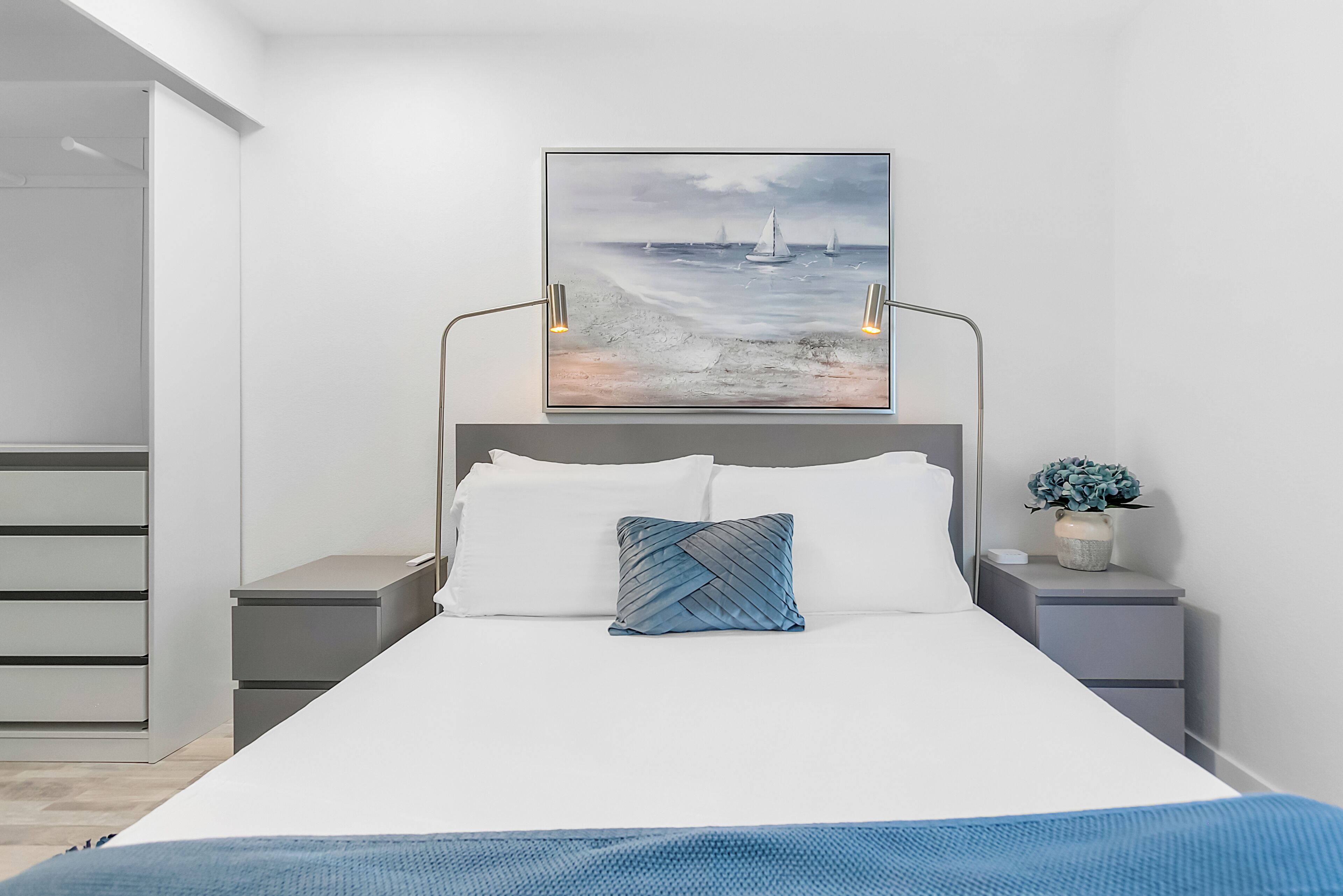 A modern bedroom with a large bed, a headboard with a painting, bedside tables with lamps, and a small plant.