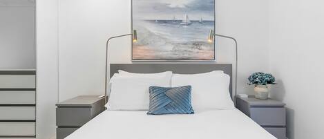 A modern bedroom with a large bed, a headboard with a painting, bedside tables with lamps, and a small plant.