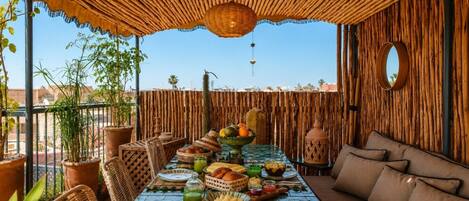 A dining area with a long table set for a meal, surrounded by wicker chairs and a sofa, with a bamboo wall and a clear view of the sky.