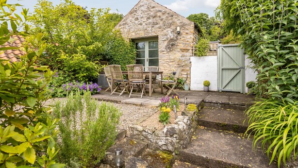 Une maison en pierre avec un petit patio, une table et des chaises, et un jardin agrémenté de diverses plantes et fleurs.