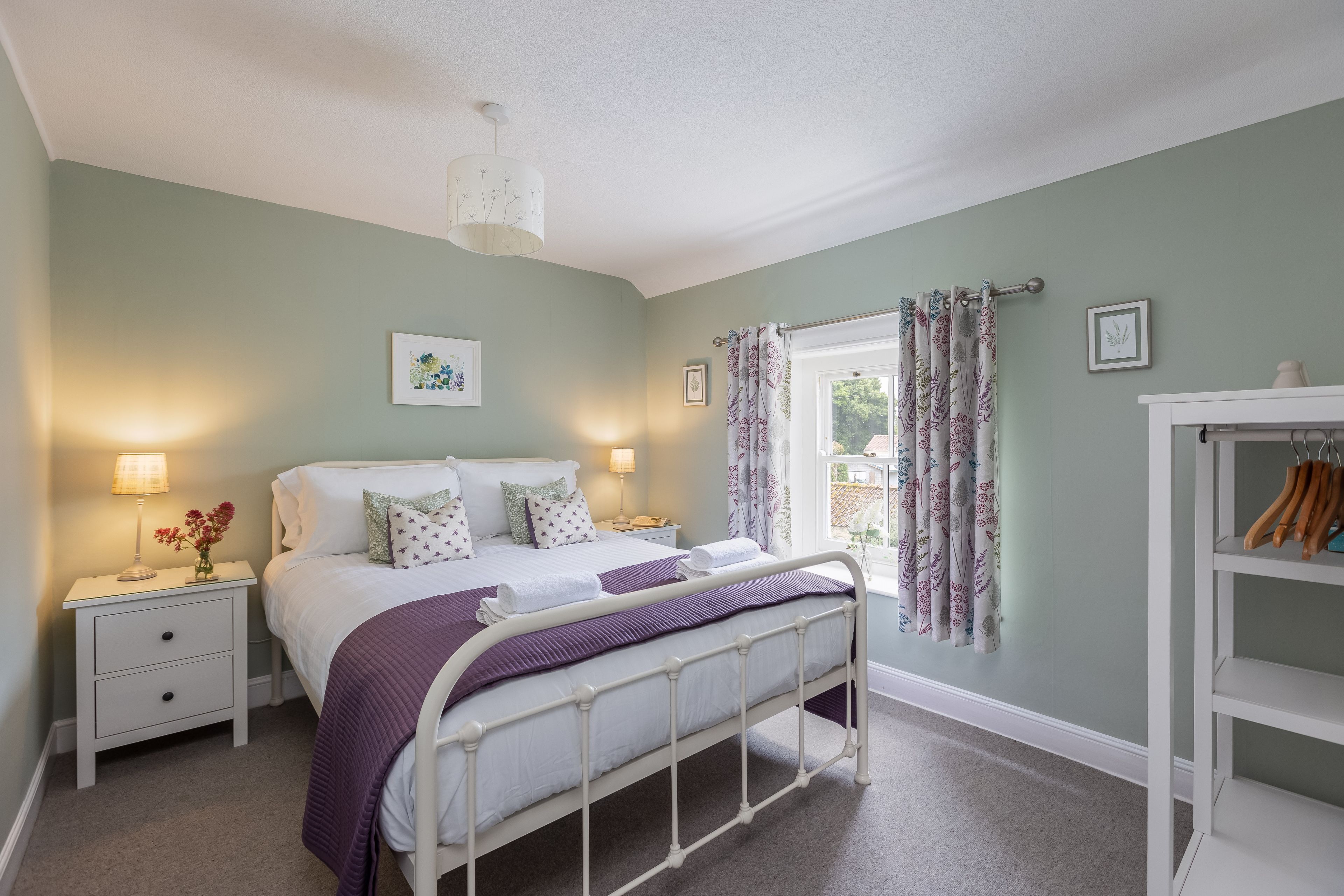 A bedroom with a bed, nightstands, a lamp, a window with curtains, and a shelf with items.