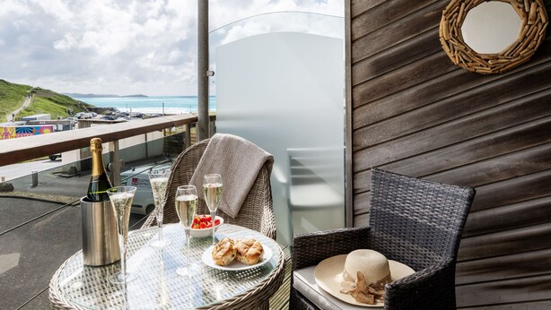 Een balkon met een rieten stoel, een tafel met champagne, glazen en gebak, met uitzicht op een strand en een parkeerplaats.