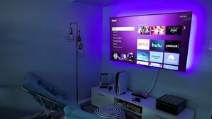 Ein Zimmer mit einem großen, an der Wand montierten Flachbildfernseher, der eine Streaming-Dienstanwendung anzeigt. Es gibt ein Bett mit blauer Decke, einen Nachttisch mit Lampe und ein kleines Regal mit verschiedenen Gegenständen.
