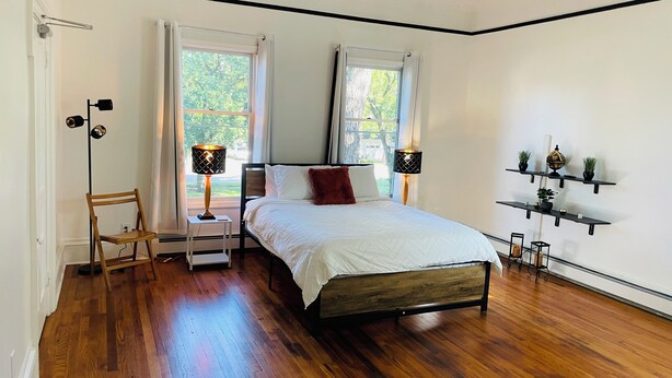 Un dormitorio con suelo de madera, una cama con ropa de cama blanca, dos lámparas de noche, una silla y un estante con objetos decorativos.