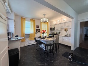 A modern kitchen with white cabinets, a dining area with a marble table, and a black dog on a rug.