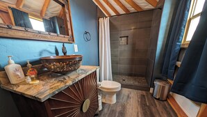 Un baño con tocador de madera, un espejo grande, un lavabo con un recipiente de mosaico, un inodoro y una zona de ducha con cortina.