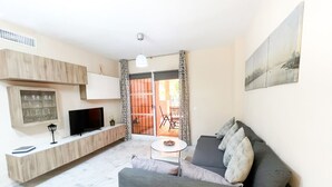 Un salon moderne avec un canapé gris, un téléviseur posé sur une console en bois et un coin repas comprenant une table et des chaises.