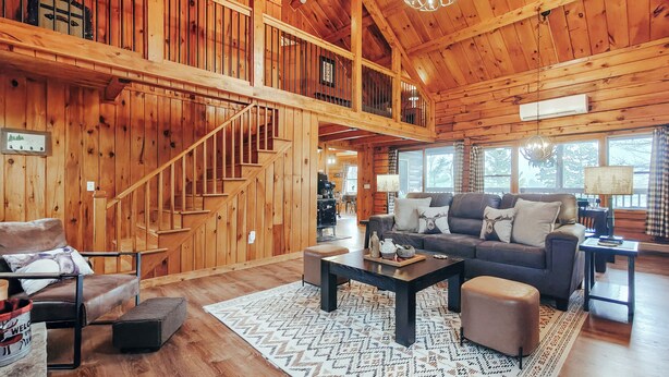 Una cabaña de madera espaciosa con una escalera que conduce a un nivel superior, una zona de estar acogedora con sofá y mesa de centro, y amplias ventanas con vistas al exterior.