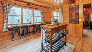 Interior de una cabaña de madera con comedor, una mesa con sillas, un ventanal con cortinas y un letrero que dice "Maine".