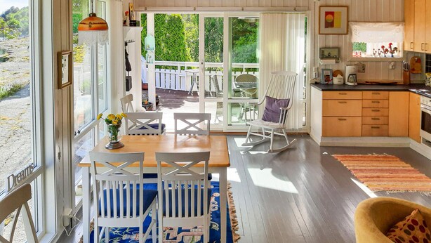 Un vaste espace de vie comprenant une table à manger en bois, des chaises blanches, un fauteuil à bascule et un tapis à motifs floraux bleus.