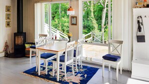Un coin repas accueillant avec une table en bois et des chaises blanches, un tapis bleu, et une vue sur une terrasse en bois et la verdure environnante.