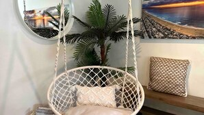 Una habitación con una silla colgante de macramé, un espejo redondo, una planta en maceta, un pouf tejido y un cuadro con una escena de playa.