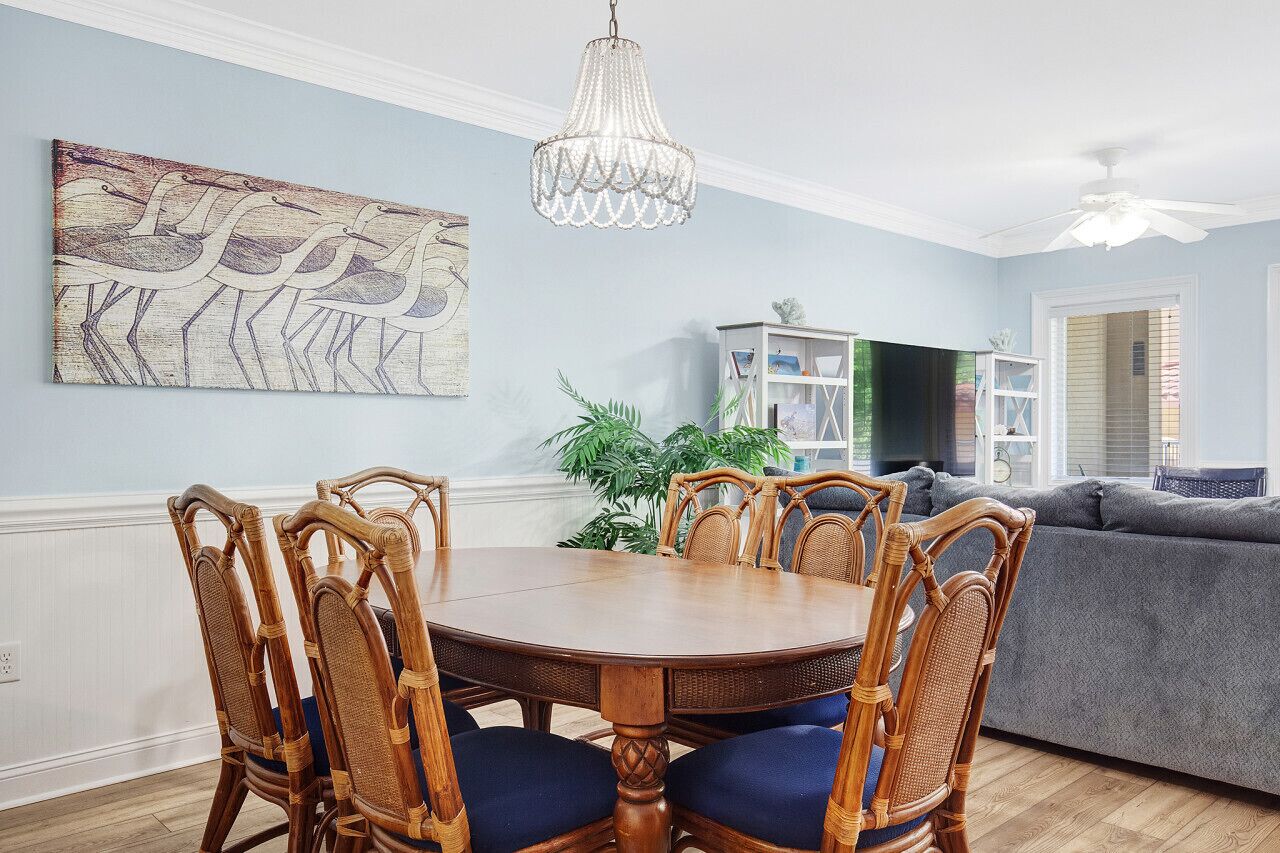 A dining area with a round wooden table and wicker chairs, a chandelier, and a painting of birds on the wall.