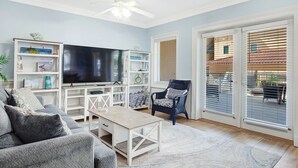 Un salon comprenant un téléviseur à écran plat, un canapé, une table basse et un fauteuil. On y trouve également une bibliothèque et une fenêtre avec des stores.