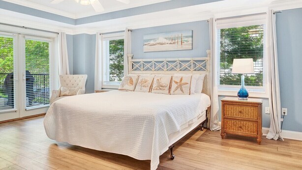 Une chambre à coucher avec un grand lit, un ventilateur de plafond, une fenêtre avec des rideaux et un tableau accroché au mur.