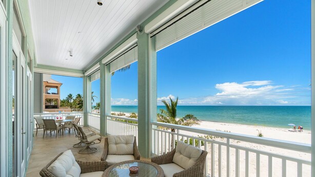 A covered outdoor seating area with wicker furniture and a glass table, overlooking a beach with palm trees and the ocean. - Biltmore Beach House | Luxury 5-Br Beachfront Pool & Spa House | Sleeps 10 (Bonita Springs)