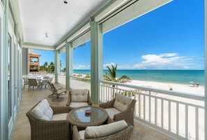Ein überdachter Außenbereich mit Korbmöbeln und einem Glastisch, mit Blick auf einen Strand mit Palmen und dem Meer.