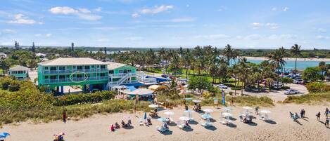 A beach resort with a large building, multiple beach umbrellas, and palm trees.