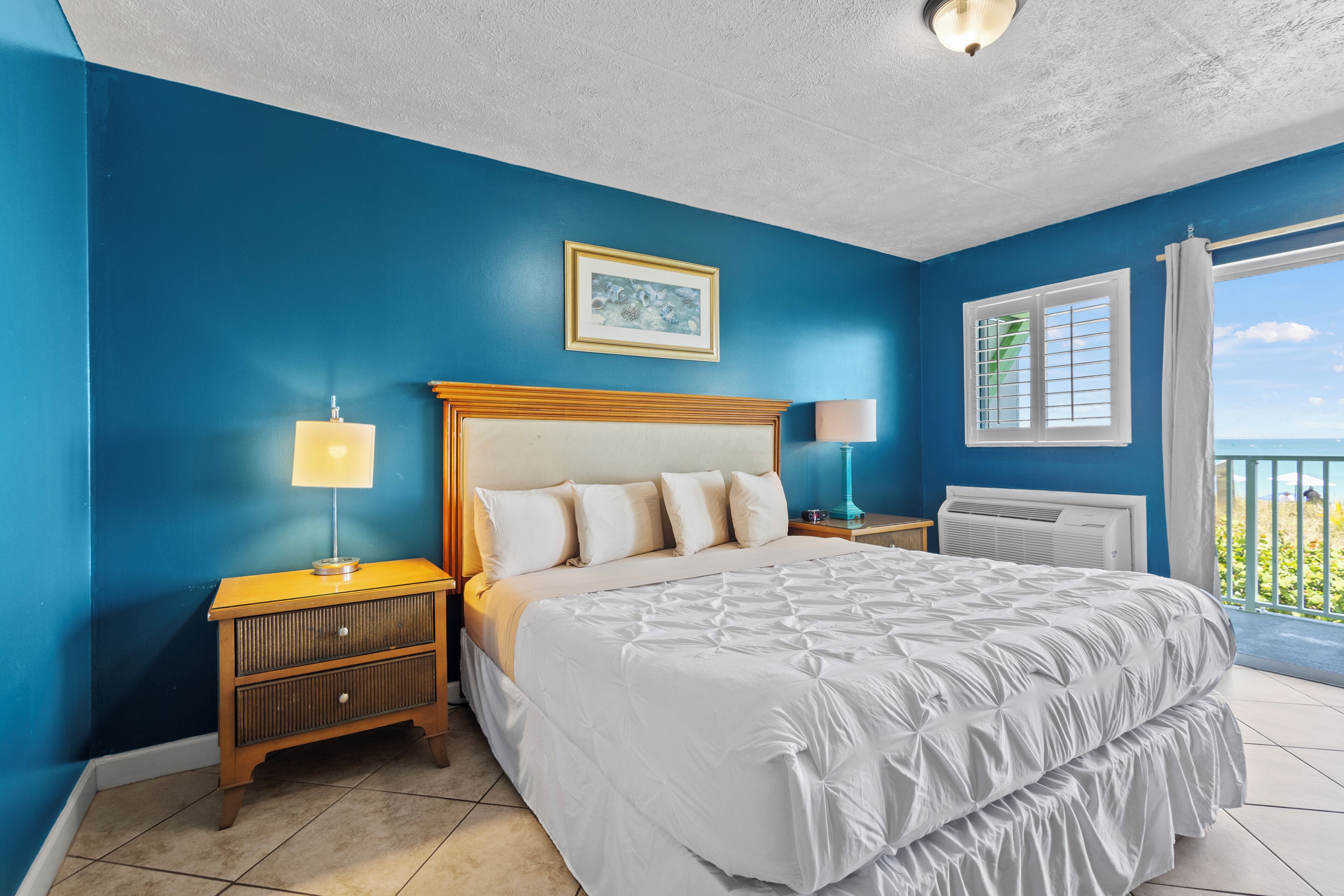 A bedroom with a large bed, two bedside tables with lamps, a ceiling light, a window with curtains, and a view of the outdoors.