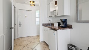 Una cocina compacta con armarios blancos, microondas, cafetera y un bote de basura.
