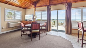 A room with a wooden ceiling, a dining area with chairs and a table, a window with blinds, and a view of the ocean. - Oceanfront Inn Suite, Ocean and Inlet Views, King Bed, #12 (Fort Pierce)