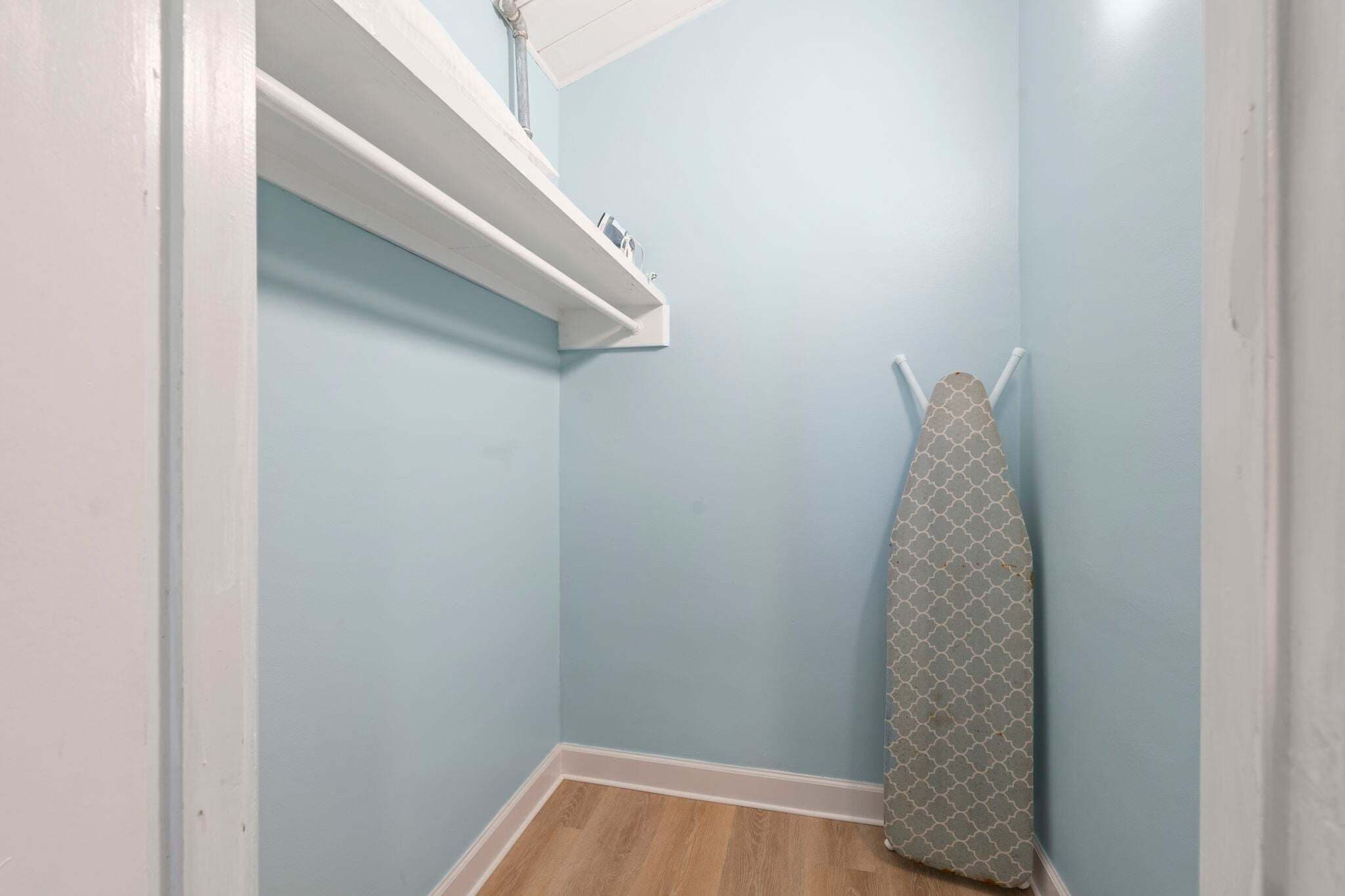 Una habitación pequeña y cerrada, con una pared azul claro, piso de madera, un estante blanco con algunos objetos, y una tabla de planchar estampada apoyada contra la pared.