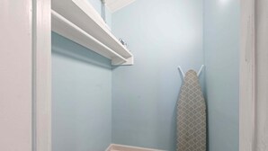A small, enclosed room with a light blue wall, a wooden floor, a white shelf with a few items, and a patterned ironing board leaning against the wall. - Oceanfront Inn, Standard Room, King Bed, Town View, #11 (Fort Pierce)