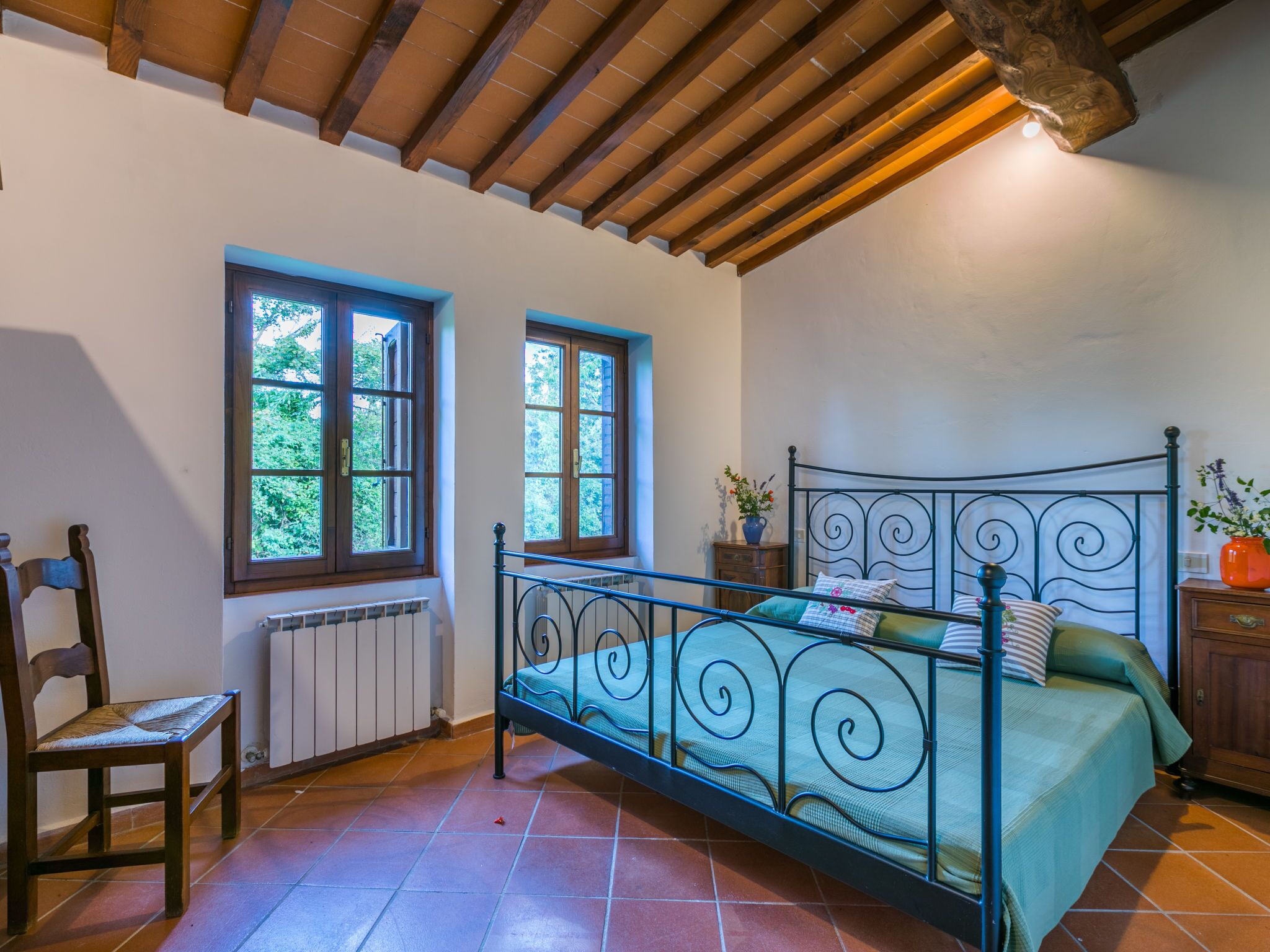 A bedroom with a metal bed frame, two windows, a radiator, and a wooden chair.