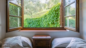 Une chambre avec une grande fenêtre donnant sur un jardin luxuriant et verdoyant. On y trouve deux lits avec une literie à motifs.
