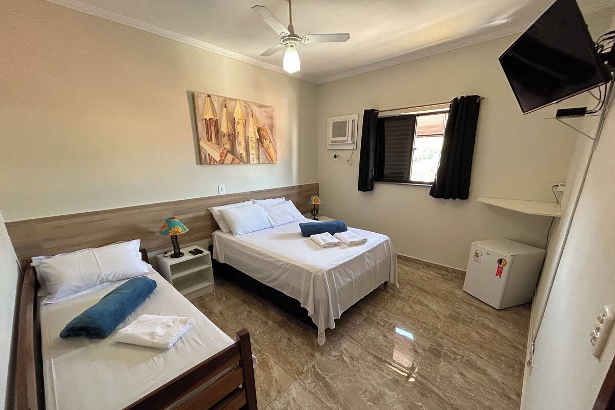 Habitación de hotel con dos camas, ventilador de techo, televisor, refrigerador pequeño y ventana con cortinas.