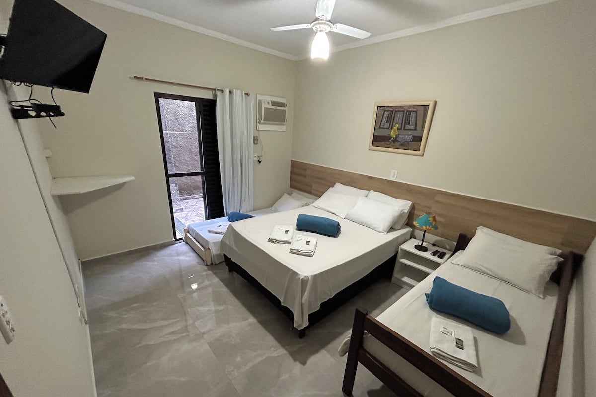 Habitación de hotel con dos camas, ventilador de techo, ventana con cortinas, equipo de aire acondicionado y un cuadro en la pared.