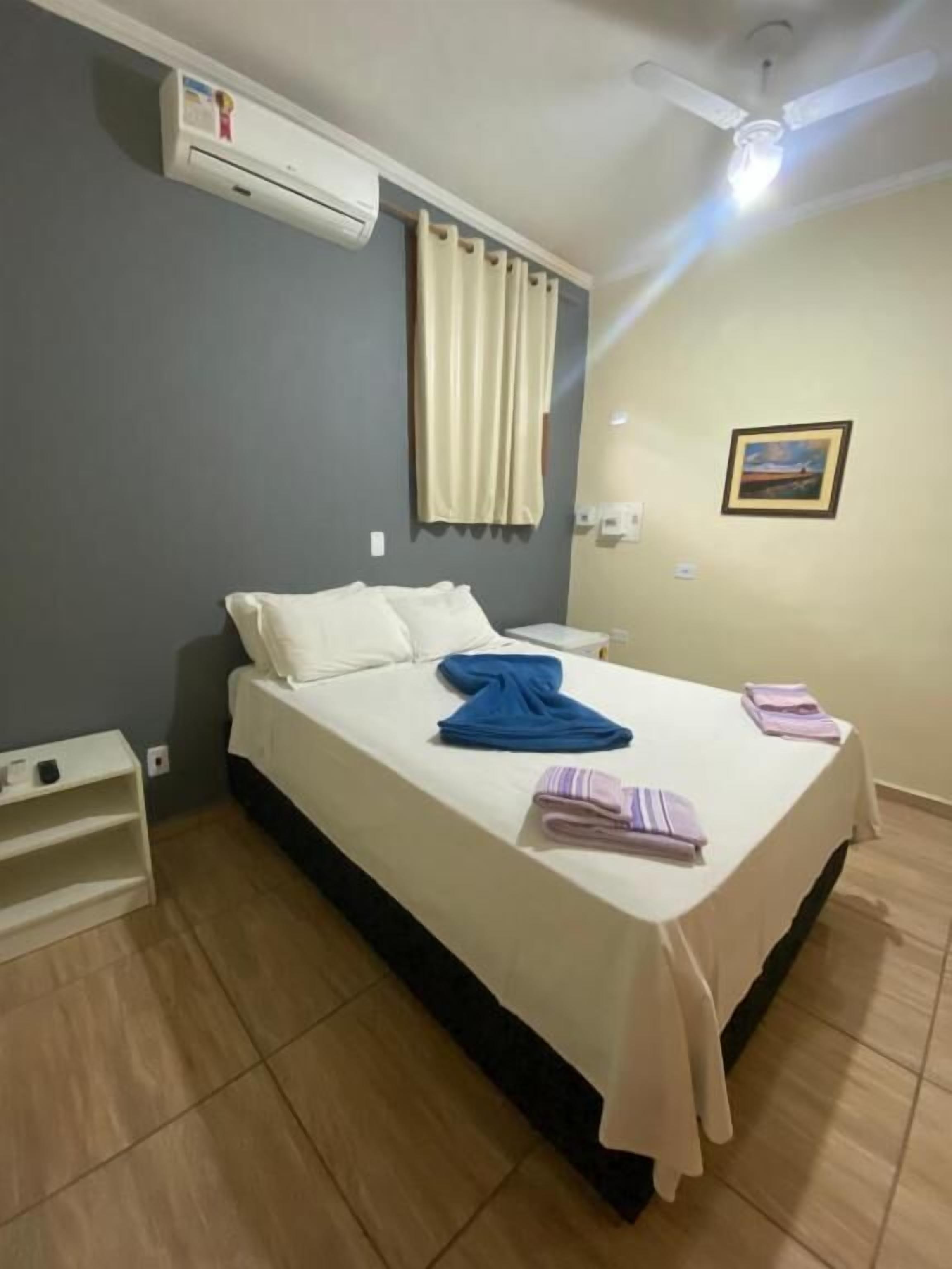 Hotelkamer met een bed, plafondventilator, airconditioning, nachtkastje en een schilderij aan de muur.
