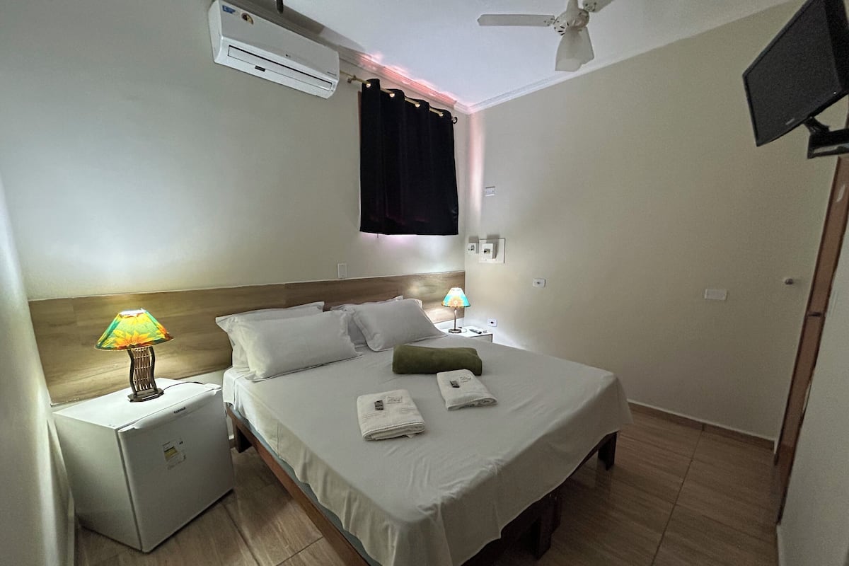 Habitación de hotel con cama, mesitas de noche, lámpara, aire acondicionado, ventilador de techo y televisor.
