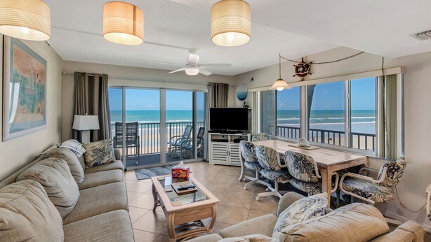 Ein geräumiges Wohnzimmer mit einer großen Couch, einem Couchtisch und einem Essbereich mit gemusterten Stühlen. Es gibt einen Fernseher, einen Deckenventilator und große Fenster mit Blick auf den Ozean.