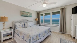 Ein Schlafzimmer mit Bett, Deckenventilator, Nachttisch mit Lampe, Kommode und Fenster mit Meerblick.