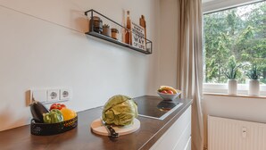 Una cocina moderna con estufa integrada, tabla de cortar con verduras, un bol de fruta y una ventana con vistas a árboles.