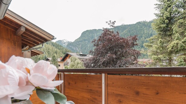 Un balcón de madera con una mesa dispuesta para dos, un florero con flores y vistas a un paisaje montañoso con árboles y un edificio al fondo.