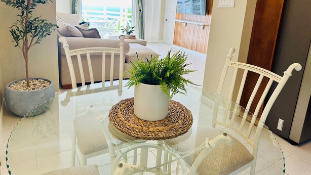 Una sala moderna con una mesa de comedor de vidrio, sillas blancas, una planta en maceta y un sofá.