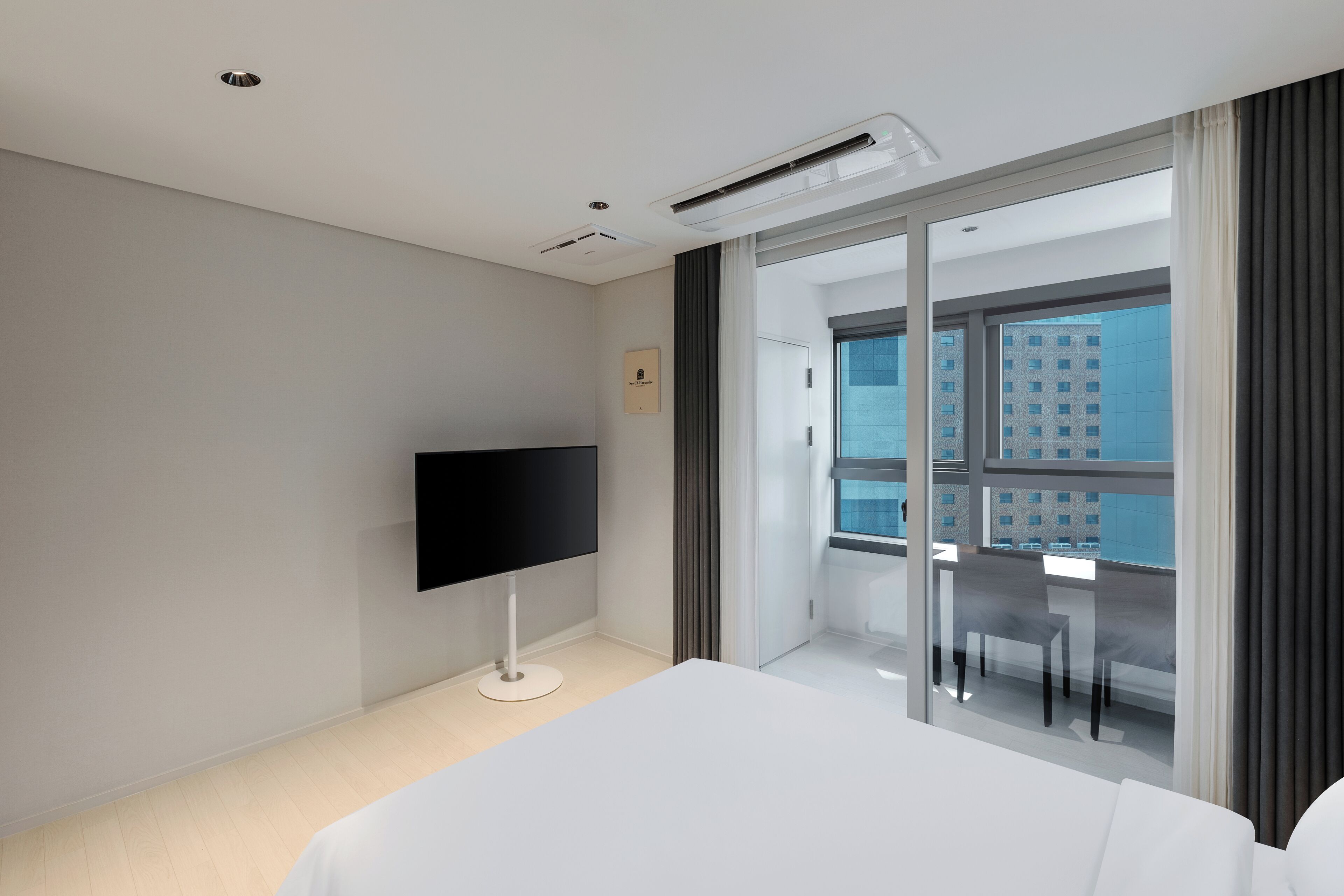 A modern hotel room with a large bed, a flat-screen TV, a balcony with a dining table and chairs, and a city view.